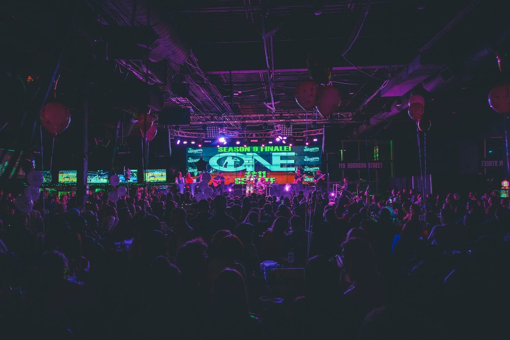 A wide view over the standing crowd toward the performance stage and lighting at 115 Bourbon Street, published on the owner’s home page. This undated concert photograph illustrates room atmosphere; it does not show a numbered seat or establish a wrestling-event floor layout.