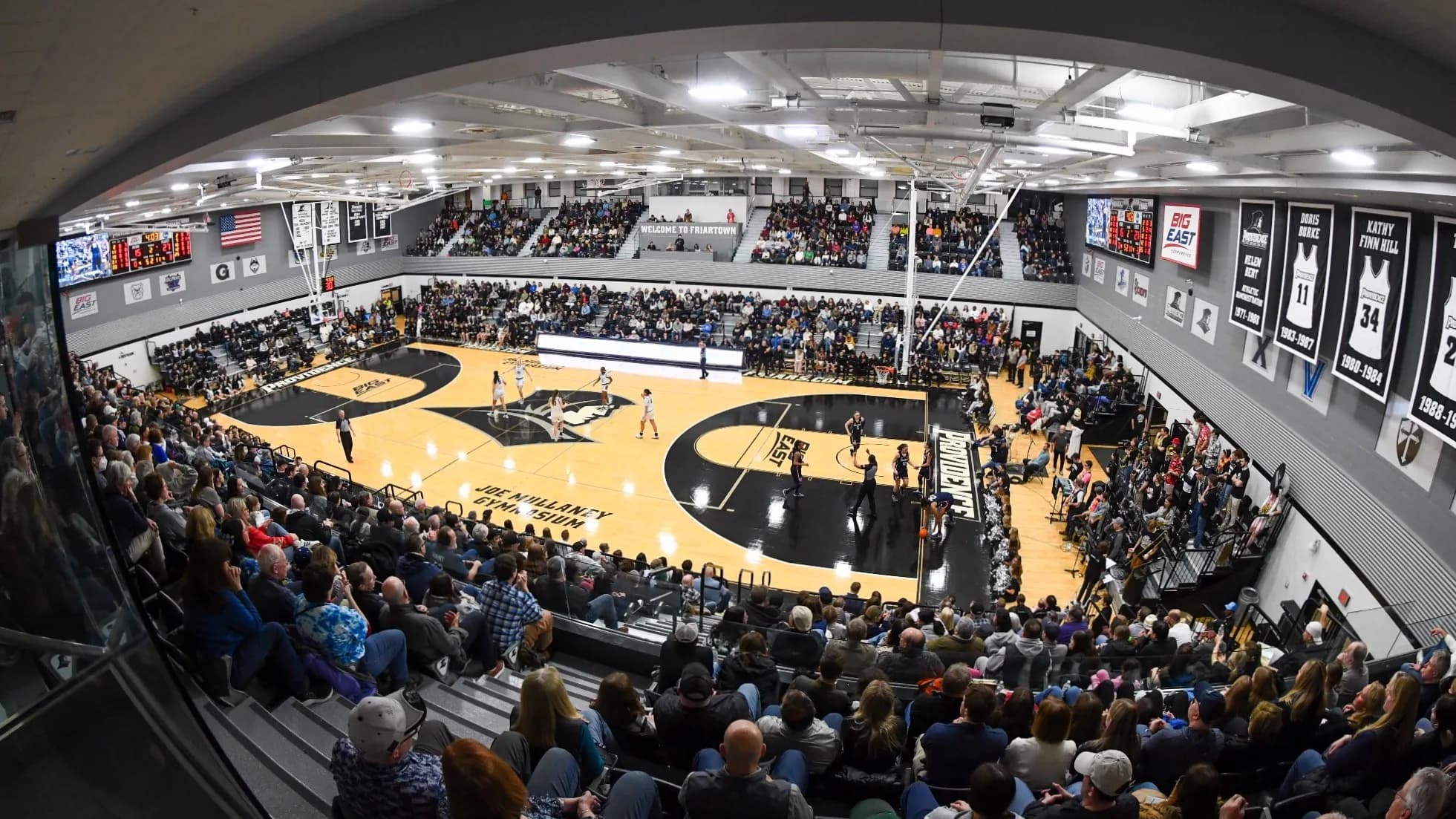 Alumni Hall/Mullaney Gymnasium during Providence–UConn on March 2, 2024, in Erica Denhoff’s photograph credited by Providence to Icon Sportswire. The elevated corner view shows both seating levels, the court, stepped aisles and railings; spectators and the near basket affect sightlines. This historical game photograph does not identify an exact ticketed seat.