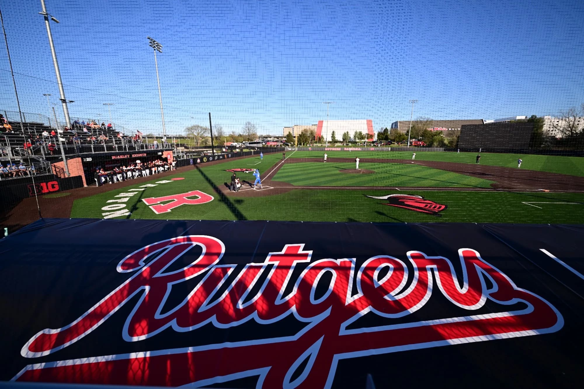 Bainton Field’s diamond and outfield seen through protective netting in Rutgers Athletics’ 2026 facility collection. The foreground Rutgers marking, rails and nearby stadium structures remain visible. No numbered camera seat or exact capture date is supplied.