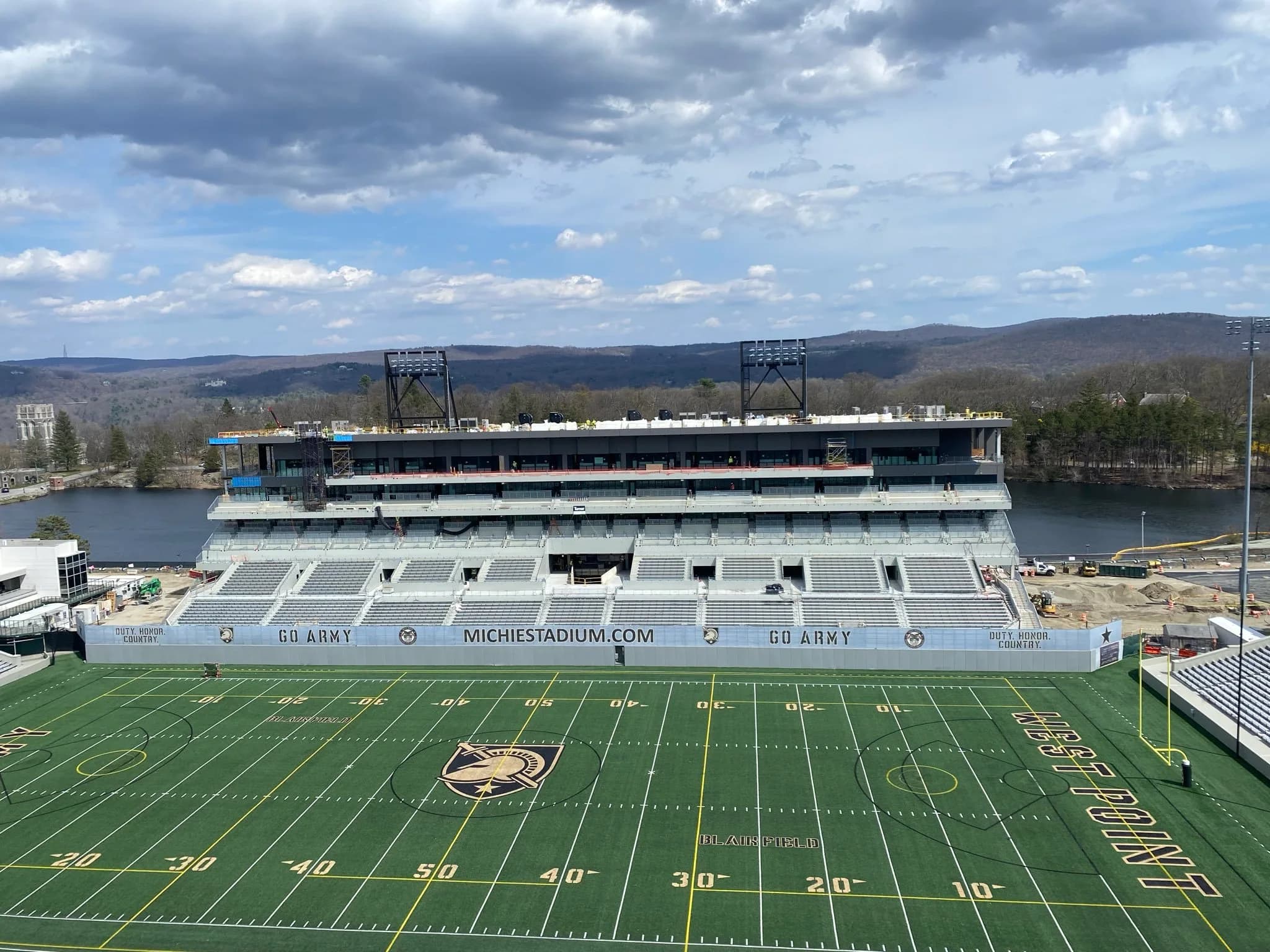 Construction-stage view across Blaik Field toward Michie Stadium’s rebuilt East Stands, showing the new cadet seating and premium structure with Lusk Reservoir behind. The owner project page supplies this actual progress photograph; scaffolding and unfinished areas are visible. It is not a rendering or a photograph of the final game-day layout, and the exact capture date is unstated.