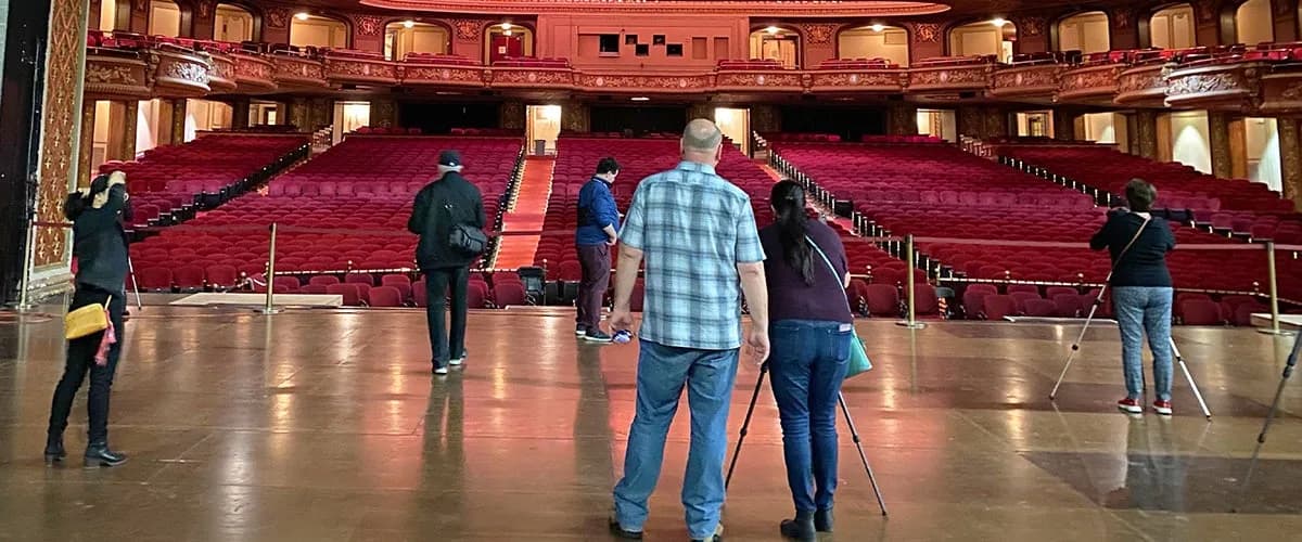 Visitors on the Wang Theatre stage during a tour, facing the red orchestra seats and ornate balcony fronts. This official tour photograph does not imply stage access at a performance; capture date is unspecified.