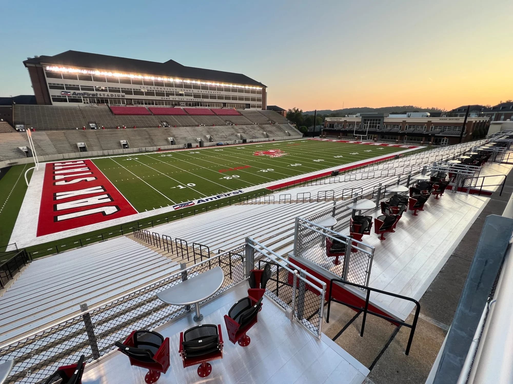 AmFirst Stadium’s field, opposite grandstand and premium tabletop seats at sunset, with the west-end football performance building beyond. Owner facility photograph published in the 2024 gallery; exact capture date is unstated.