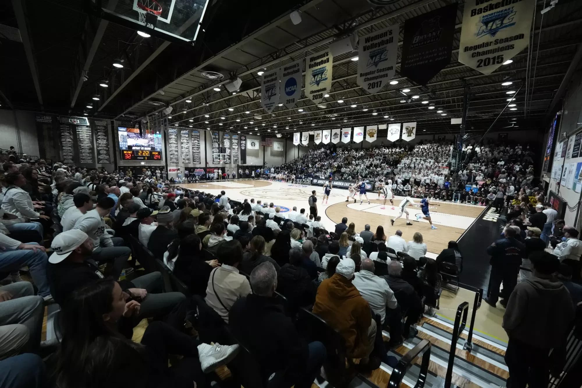 Chace Athletic Center’s actual basketball court and occupied seating from a corner in Bryant’s official facilities collection. Standing spectators, the near basket and aisle railings affect the view. Capture date and individual ticketed-seat position are not supplied.