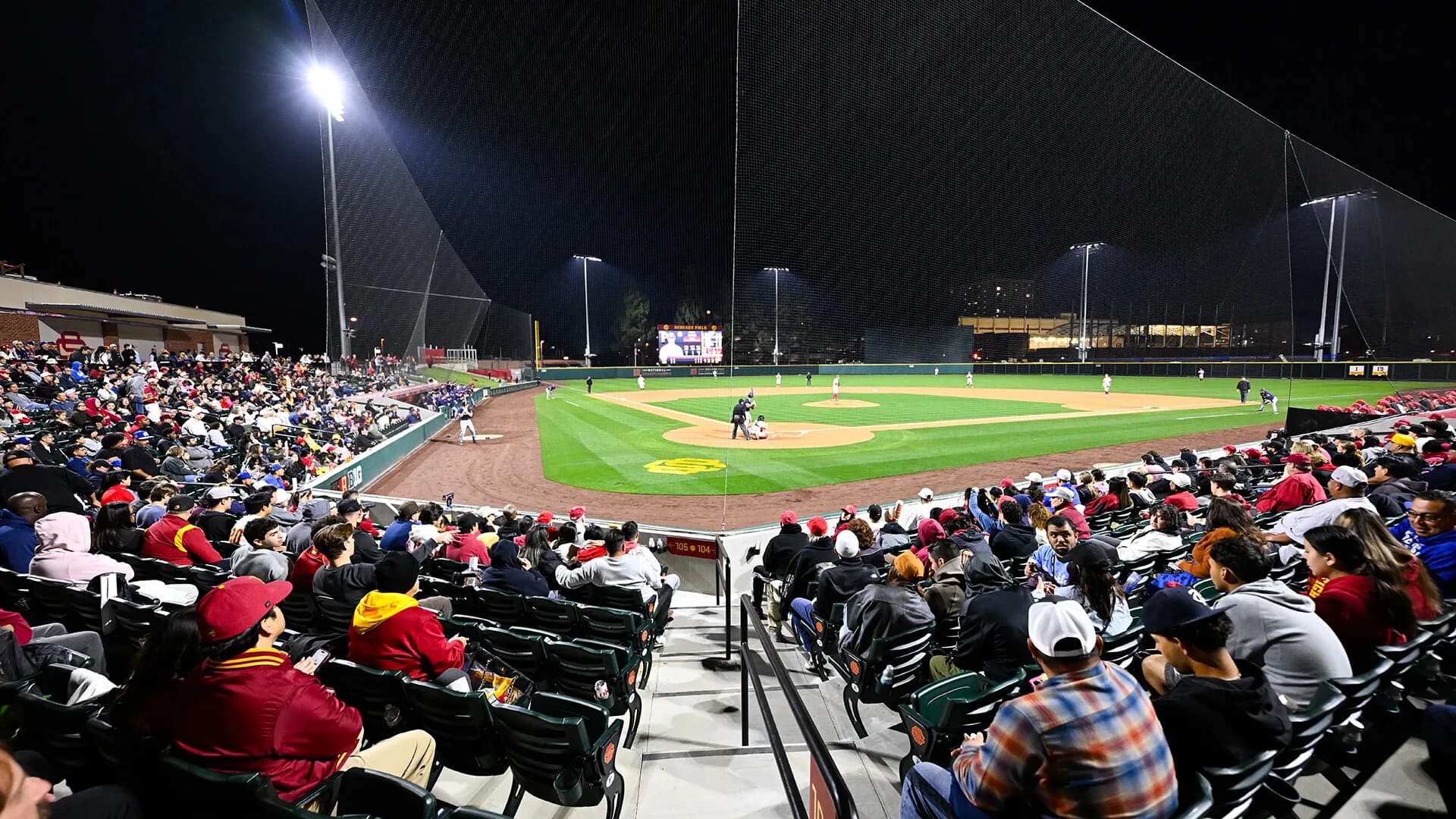 The rebuilt Dedeaux Field’s occupied bowl and diamond at night in USC Athletics’ 2026 facility collection. Nearby spectators, aisle railings and protective netting remain visible. No numbered camera seat or exact capture date is supplied; compare the actual game’s setup.