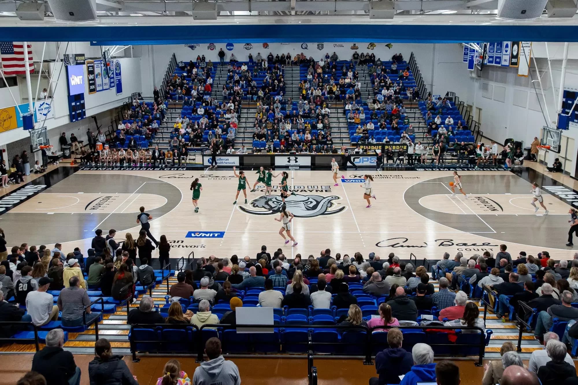 Gates Sports Center’s basketball court and occupied chairbacks in Purdue Fort Wayne’s official facility photograph, with a March 2025 source path. The court, opposite seating bank and aisle railings are visible. The upload date is not independently a capture date; confirm current game seating.