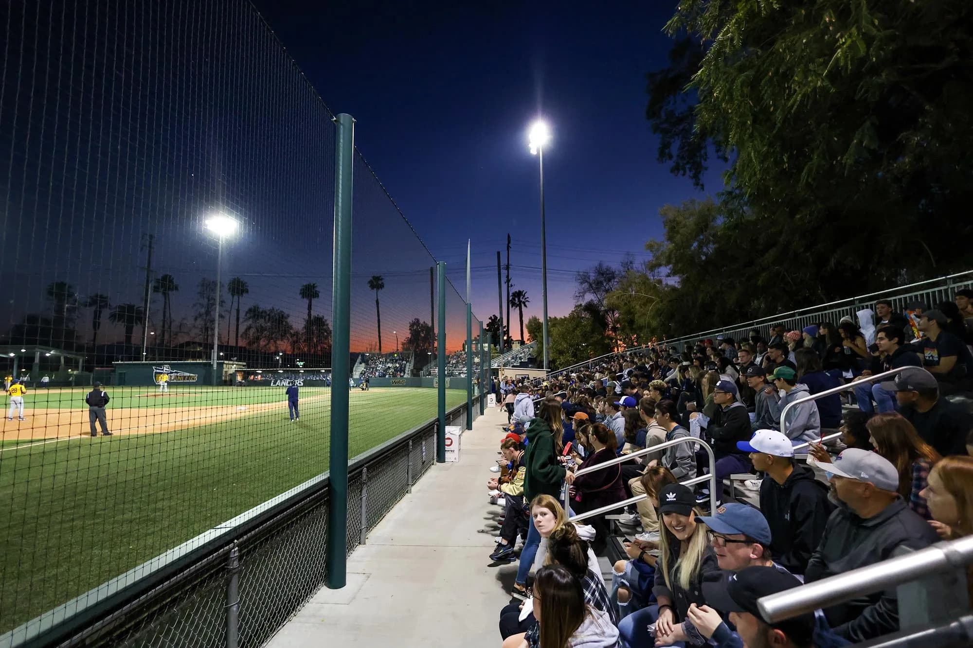 Totman Stadium’s occupied spectator seating, aisle, protective netting and illuminated diamond in California Baptist Athletics’ official collection. This actual night-game view supplies no exact capture date or numbered camera seat; retain the foreground rail and netting when judging the view.