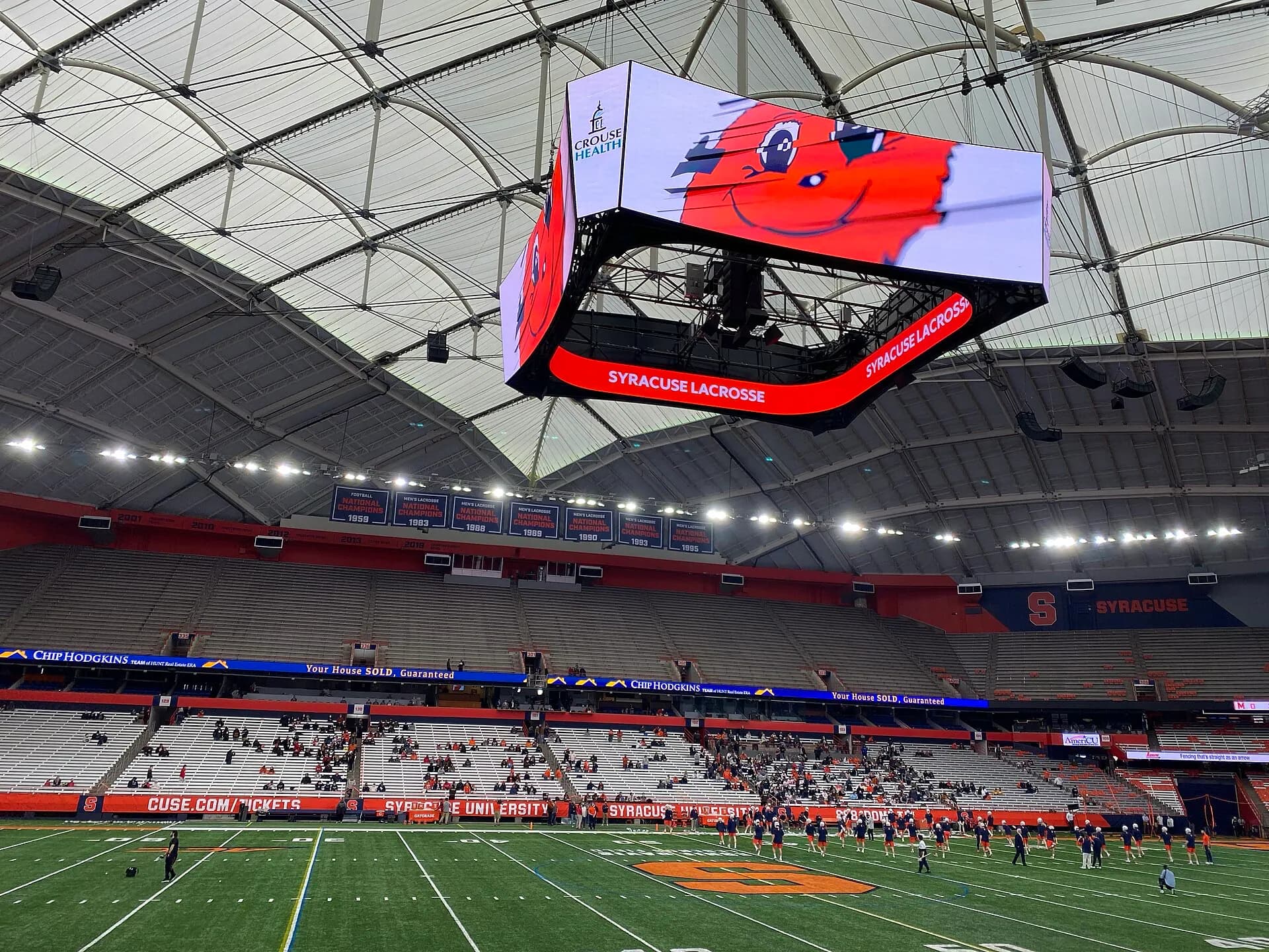 A wide interior view of the JMA Wireless Dome's field, seating bowl, video board, and roof.