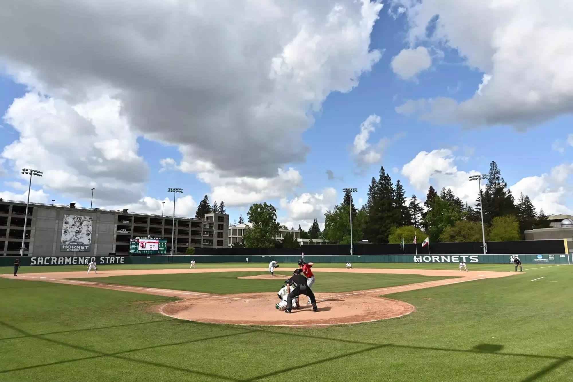 John Smith Field’s diamond and outfield seen through protective netting from behind home plate in Sacramento State Athletics’ earlier-season facility collection. Poles, foreground netting and the batter/umpire remain visible. No exact capture date or numbered spectator camera seat is supplied.