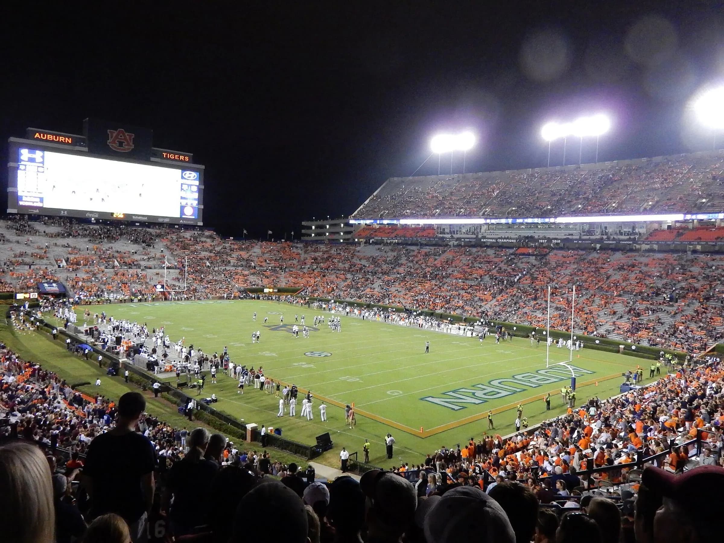 Nighttime fan view across Jordan-Hare Stadium's field, videoboard, and seating bowl.