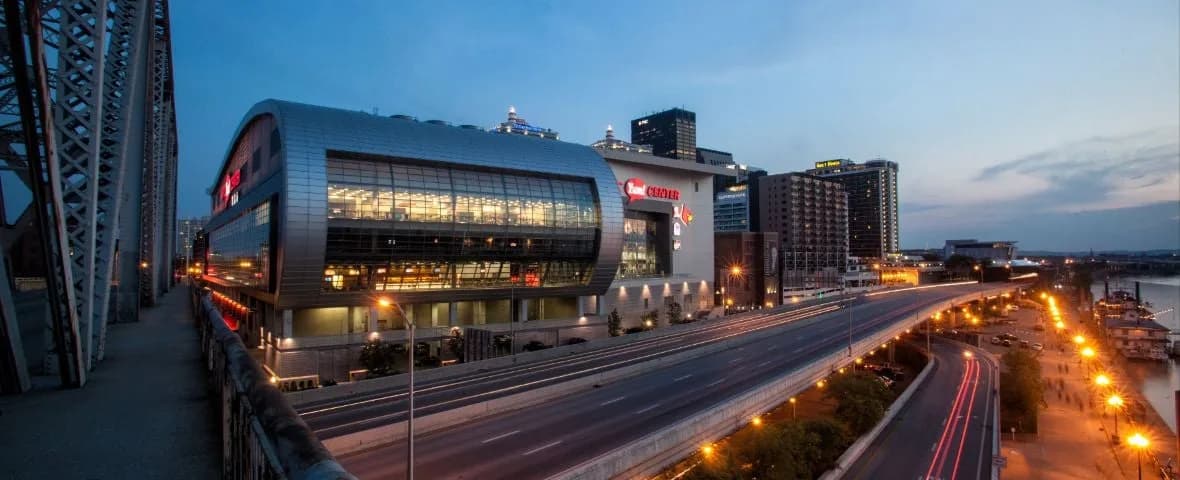 KFC Yum! Center beside Interstate 64 and the Ohio River at dusk, showing the arena’s curved glass exterior and surrounding roads. From the current official directions page; capture date is unspecified and this photograph does not establish a pedestrian route.
