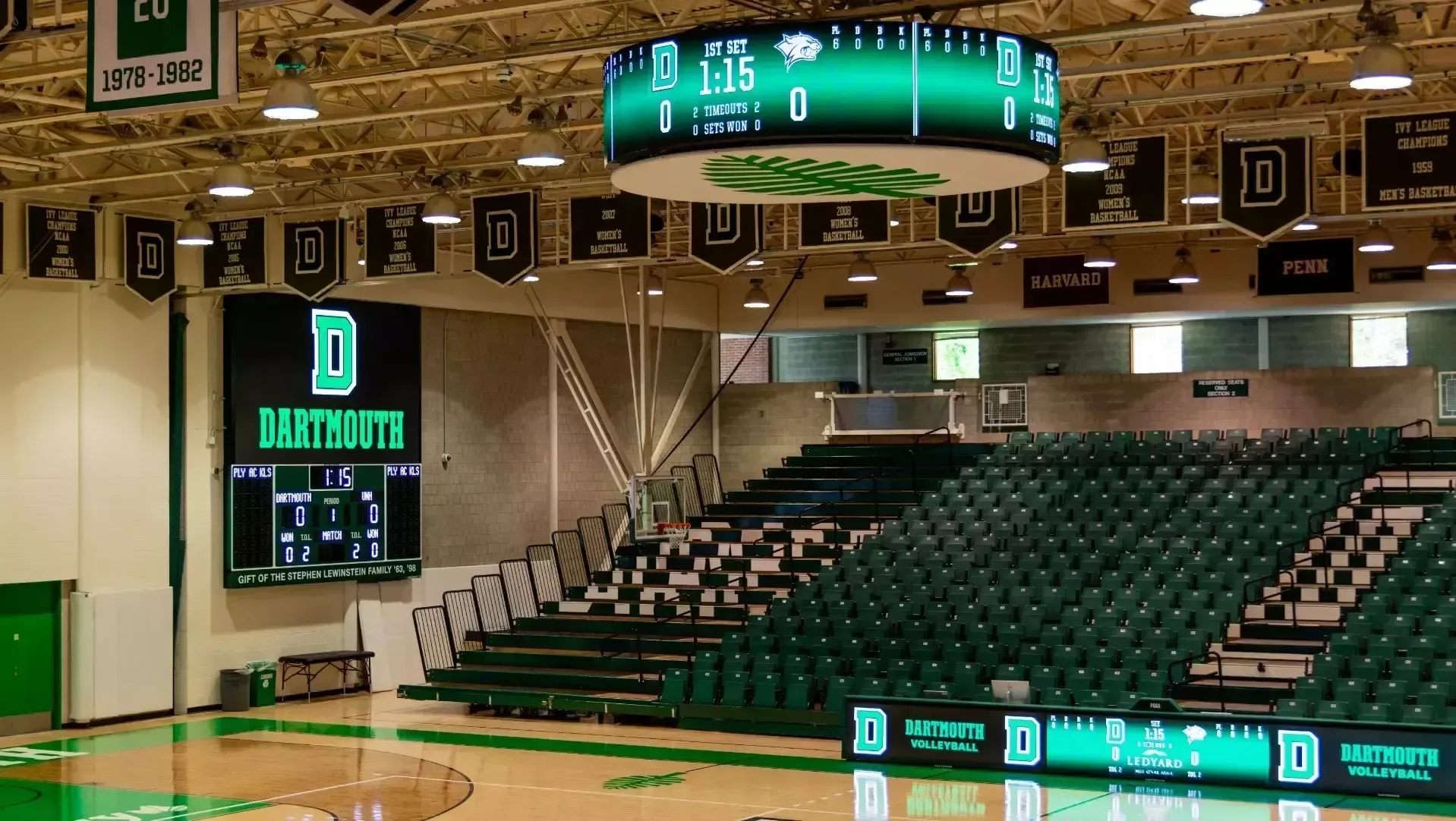 Leede Arena’s seating and video boards in Dartmouth’s September 2024-source facility collection. The image shows green chairbacks, adjoining bleachers, aisles and the center-hung display in a volleyball configuration. Basketball equipment and event seating can differ; capture date is unverified.