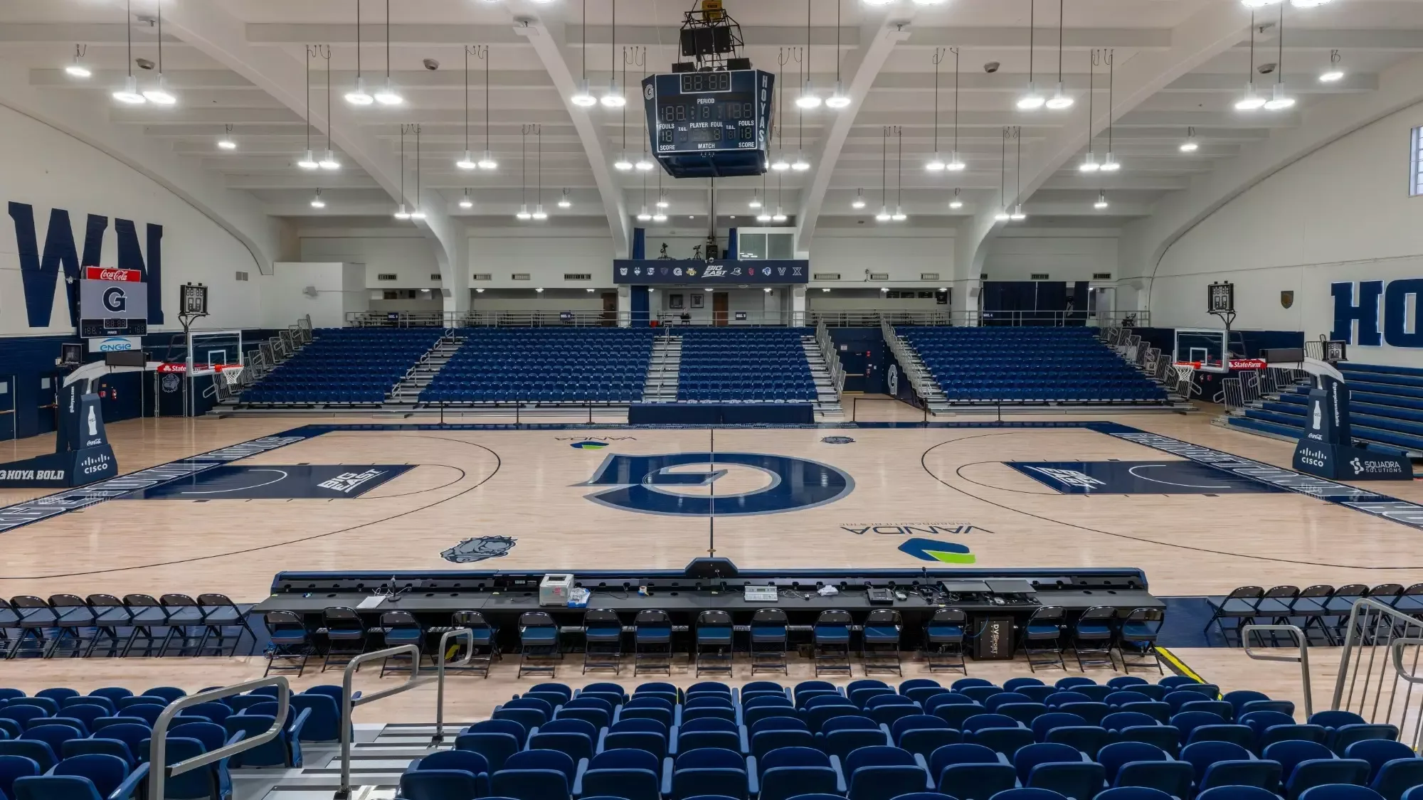 McDonough Arena’s basketball court and blue chairbacks in Georgetown’s current official facility photograph. The facility page describes the 2025 seating renovation; the October 2026 asset upload does not independently establish its capture date. Stepped aisles and foreground railings affect the view. Confirm your current event seating and accessible route.