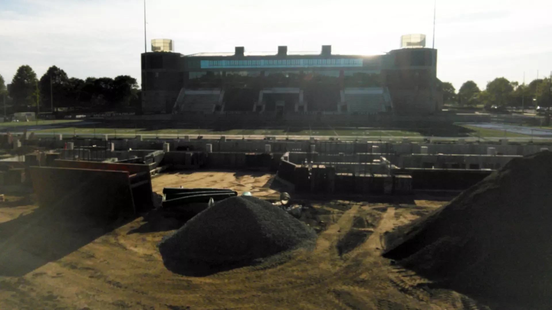 Meade Stadium’s actual east-stand construction and foundations with the Ryan Center opposite, as shown in URI’s current project update reviewed October 16, 2026. No exact capture day is supplied. The former east grandstand was demolished in March 2026; this is a worksite, not an operating spectator area.