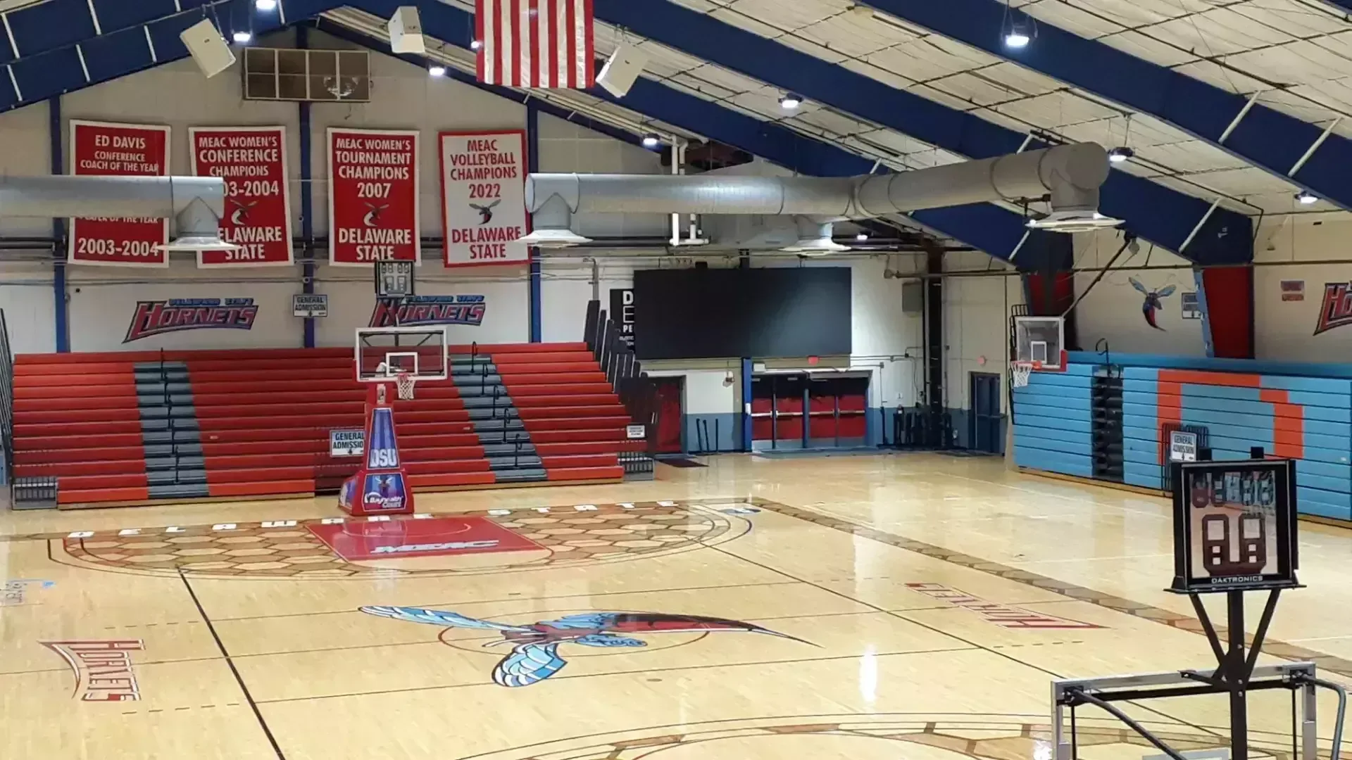 Memorial Hall / Bayhealth Court in Delaware State’s official March 2025-source facility photograph. The empty basketball floor, wall hoops and folded red-and-blue seating are visible. Retracted bleachers describe a non-event setup and do not establish the current game-day seating allocation or an exact seat view.