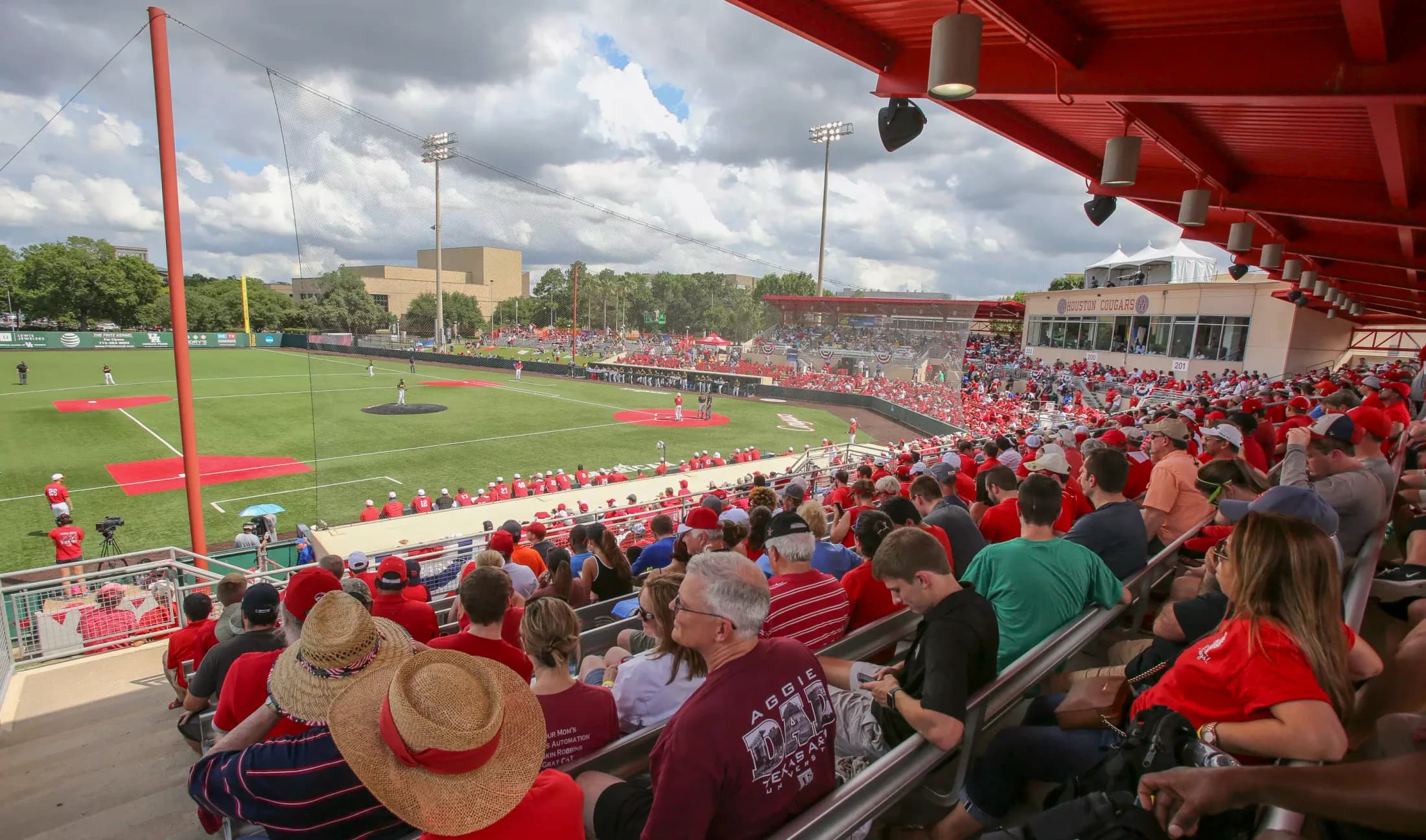 Schroeder Park’s occupied covered seating and diamond in Houston Athletics’ historical facility gallery, published June 29, 2017. Roof supports, netting, nearby spectators and railings remain visible. The source supplies no numbered camera seat or exact capture date; compare the current ticket and layout.