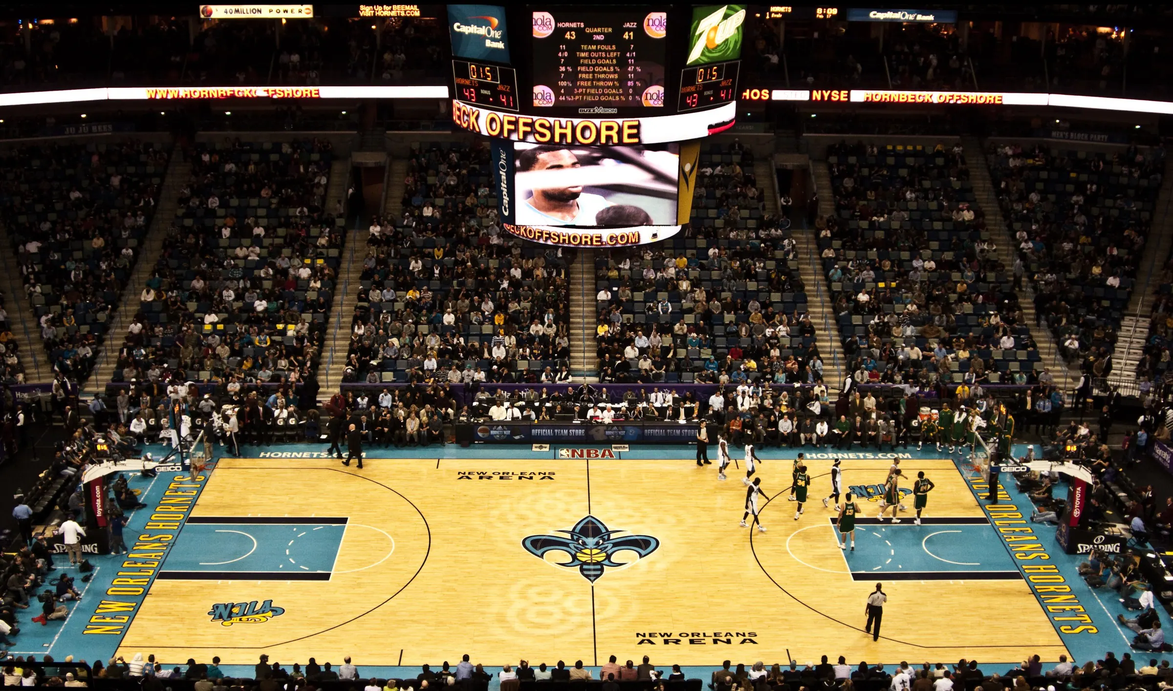 Wide centered view of Smoothie King Center's full basketball court, scoreboard, lower seating bowl, aisles, and crowd.