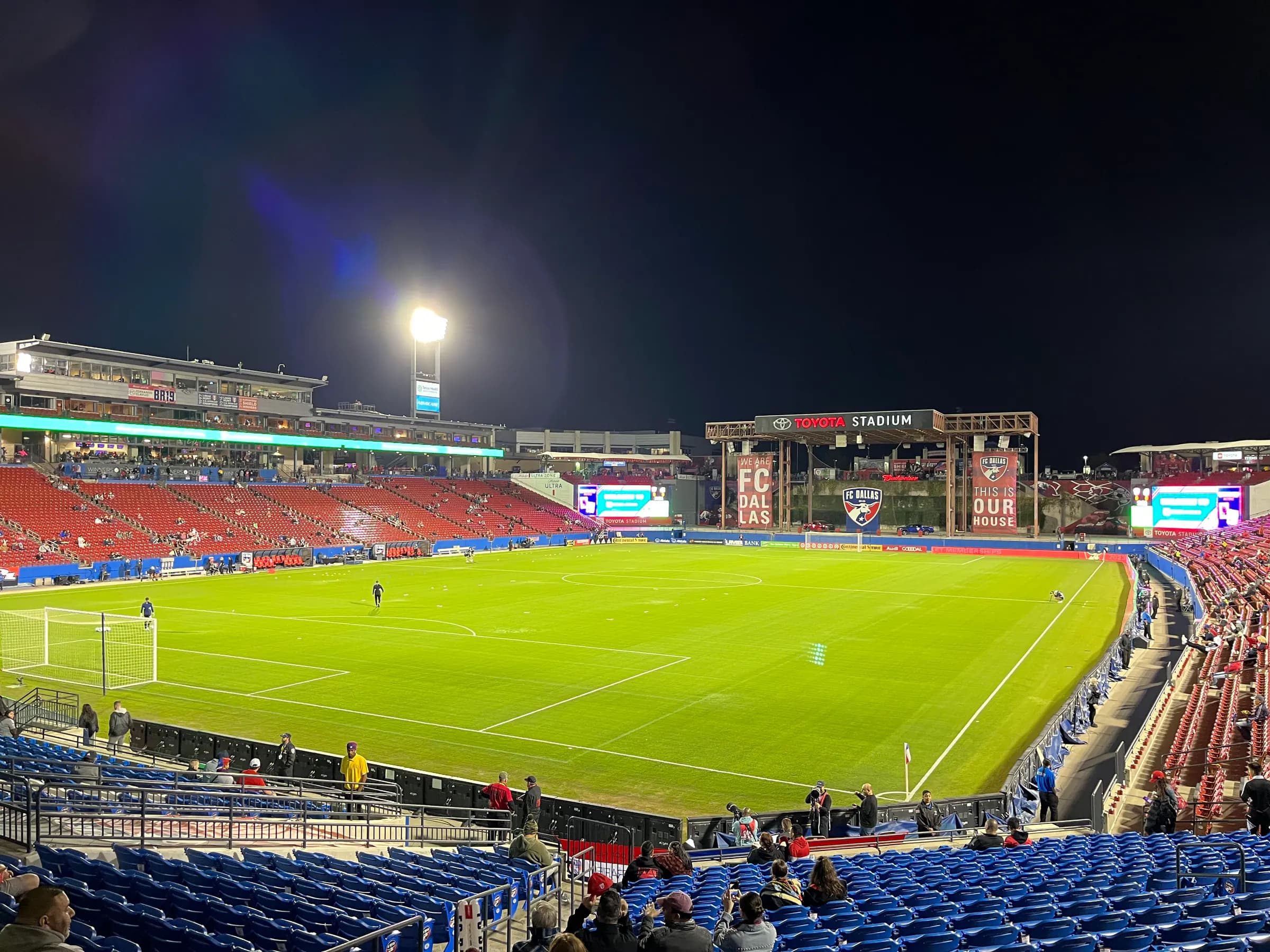 Wide fan-seat view across Toyota Stadium's full pitch, seating, video boards, and west-end entrance during a night match.