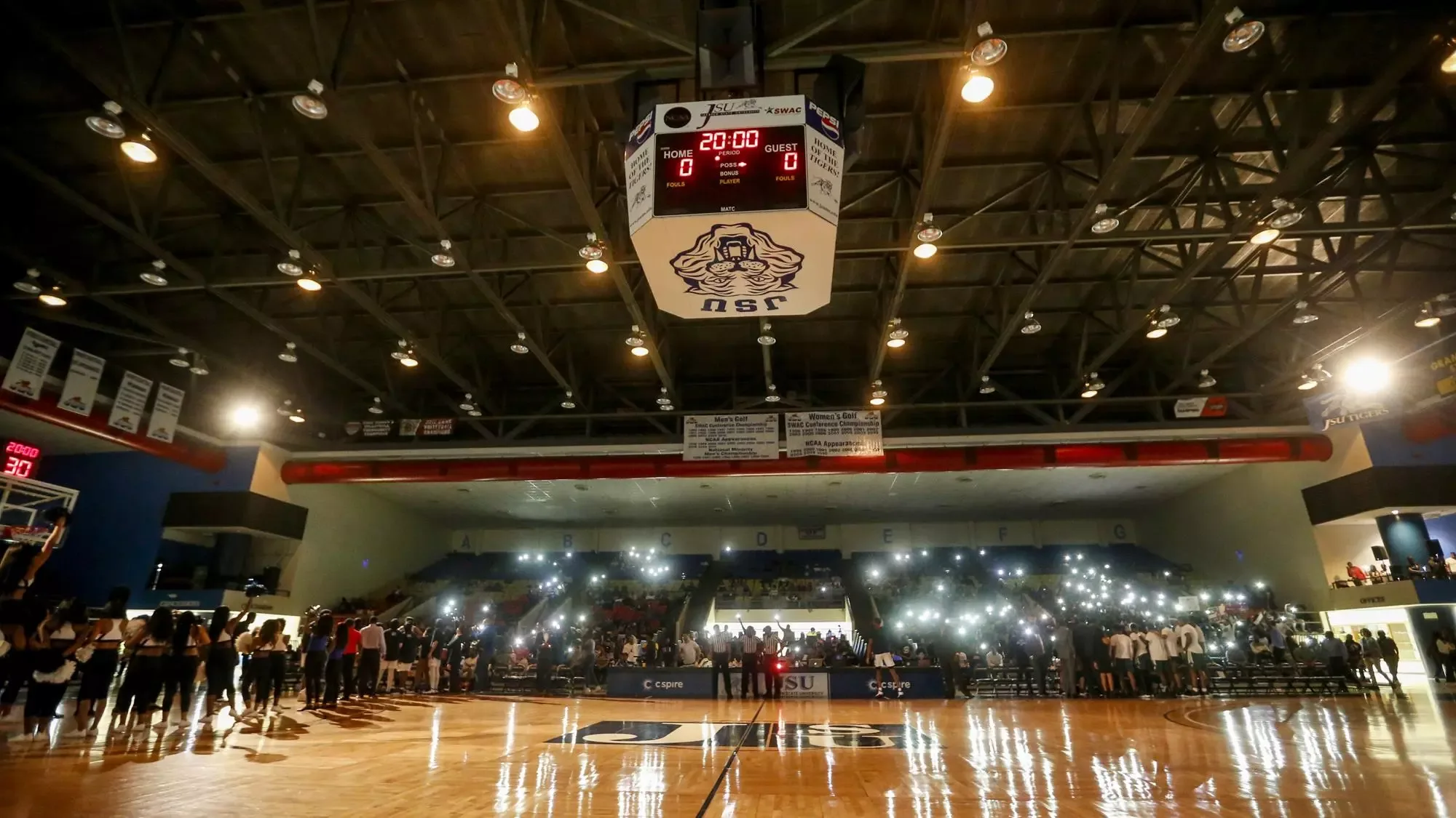 Jackson State’s home basketball arena during pregame lighting in the official facility collection, with an October 2019 source path. The JSU scoreboard, blue chairs, grey bleachers and lettered sections identify the bowl. This court-level view does not identify a ticketed seat or establish today’s allocations.