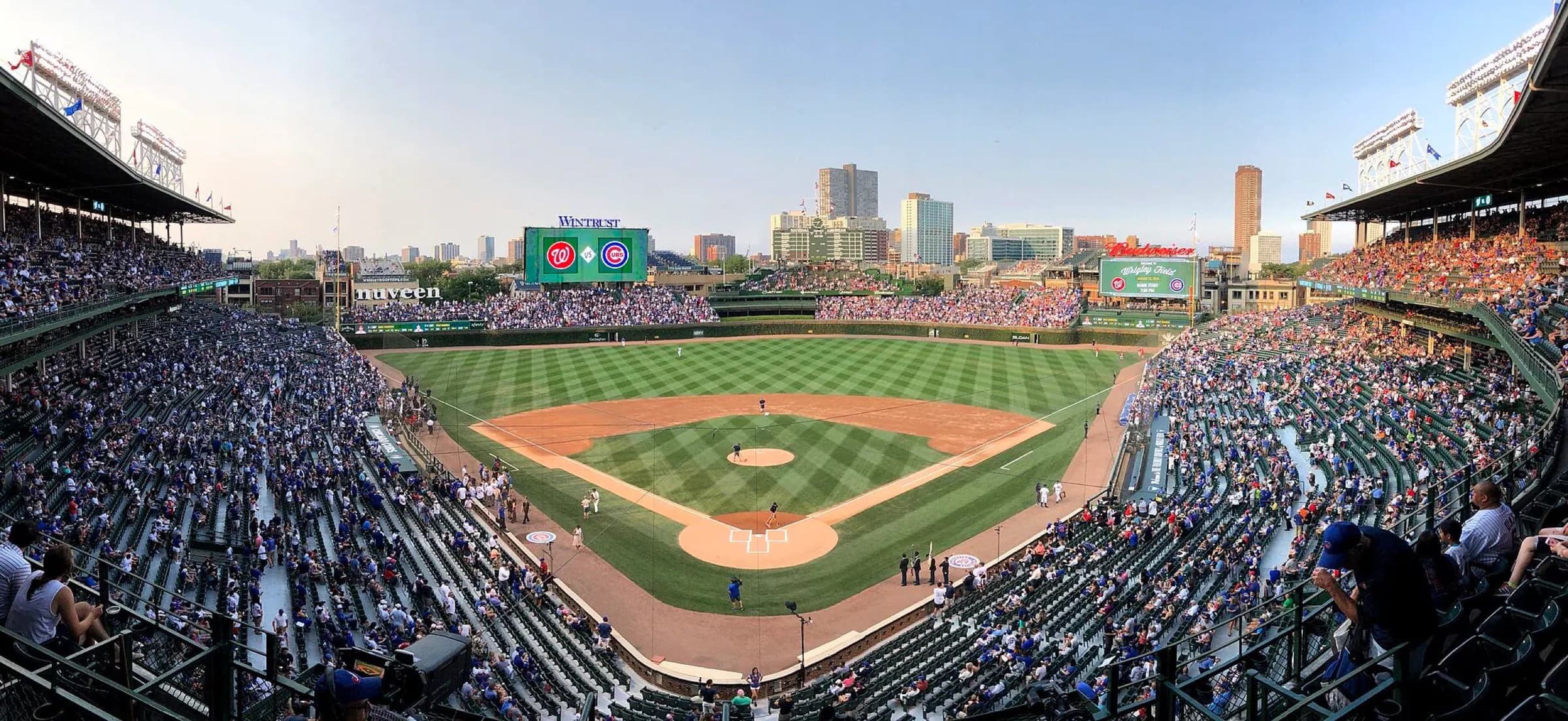 Panoramic upper-deck view from behind home plate across Wrigley Field's diamond, seating bowl, outfield bleachers, scoreboards, and neighborhood skyline.